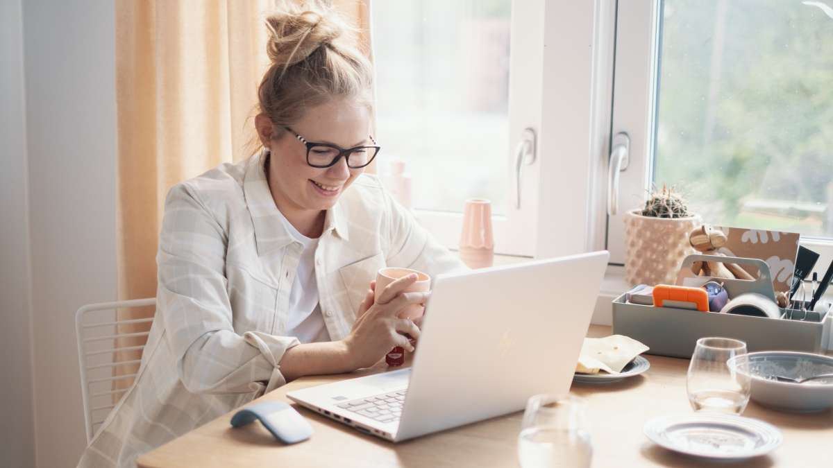 Kvinne sitter ved vindu, og smiler foran sin laptop.